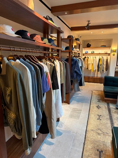 The image captures the interior of a retail space, seemingly a clothing store, with racks of apparel neatly displayed on wooden shelves and hangers. Predominantly, the focus is on the clothing, with a variety of colors and fabrics suggesting a range of styles. The store's interior design incorporates wooden fixtures and a neutral color palette, creating a warm and inviting atmosphere. In the background, there's a mirror reflecting additional clothing displays and a glimpse of seating. The flooring appears to be a light-colored tile, and a patterned rug adds texture and visual interest to the space. Given the upscale and curated aesthetic of the store, it's plausible to infer that the location could be in West Hollywood, USA, which is known for its boutique shops and design-forward retail environments.