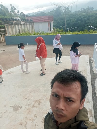 The image depicts an outdoor scene in Pangalengan, Indonesia, featuring a group of people on a concrete-paved area, likely a viewing platform or terrace, under an overcast sky. In the foreground, a man takes a selfie, his face occupying the lower right portion of the frame, looking directly into the camera with a focused expression. He is wearing a dark, possibly olive green, jacket.
Behind him, four females are visible, along with a partially seen child on the far left. A young girl in a white t-shirt and light-colored pants, wearing glasses, stands to the left, seemingly interacting with a woman next to her. This woman wears an orange hooded jacket and light pants, with black sandals. Further to the right, another woman, dressed in a light-colored hijab, white long-sleeved top, black skirt over leggings, and white sneakers, is engrossed in her phone, holding a white bag. On the far right, a young girl in a black hijab and pink top with striped pink pants is holding a small, colorful puzzle cube.
The background reveals a picturesque natural landscape dominated by lush green, forested hills and mountains, suggesting a serene, possibly rural or tourist-oriented location. A light blue barrier separates the immediate concrete area from the distant scenery. A small white building with a red roof is visible in the mid-ground, alongside a concrete retaining wall topped with several black pots. The overall impression is a casual daytime outing at a scenic spot. No visible text is present in the image.