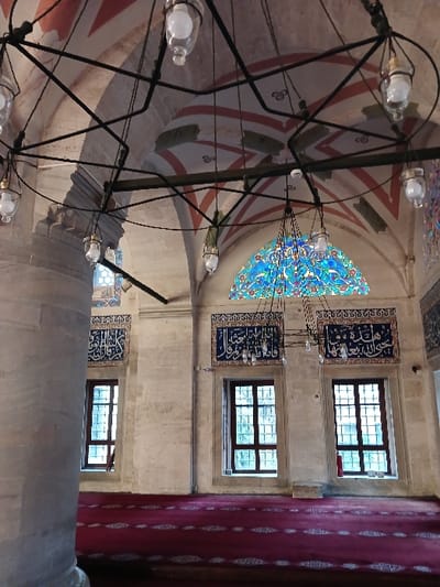 The image shows the interior of a mosque in Istanbul, Türkiye. The mosque is located at 41.025 latitude and 28.98 longitude. The scene is lit by multiple hanging chandeliers, including one large chandelier with a colorful glass shade. The ceiling has a decorative pattern painted in red and white stripes. The walls are cream-colored and adorned with blue and gold tilework featuring Arabic script. There are two large windows with wooden frames and glass panes. In front of the windows, there is a dark red carpet with an intricate pattern. The image captures the peaceful and ornate atmosphere of a place of worship.