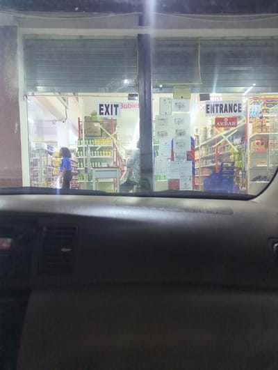 The image, taken at dusk from inside a vehicle, captures the storefront of a supermarket in Dutsen Alhaji, Nigeria. A metal roll-up door is partially raised, revealing the interior of the store which is brightly lit with fluorescent lights. Two signs are visible on the glass facade: one reads "EXIT" in large black letters on a white background, with "Babies" written in smaller pink font next to it. To the right, a larger sign displays "ENTRANCE" in bold white letters. Below the "ENTRANCE" sign is a red and white logo that reads "AKBAR PREMIUM QUALITY TEA".
Inside the supermarket, shelves stocked with various products are visible, including boxes of "CORNFLAKES". A woman in a blue shirt and dark pants stands near the left side of the entrance, facing away from the camera, appearing to be an employee. Another person, dressed in a patterned shirt, is partially visible on the right side of the store. A blue shopping cart is positioned inside the entrance. The exterior wall on the left of the building appears to be made of concrete with some textured elements. The view from the car reveals the dashboard and part of the windshield, suggesting the photo was taken while parked or slowly moving.