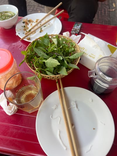The image shows a close-up, overhead view of a red plastic table set for a meal, likely in an outdoor eating establishment. In the upper left corner, a white bowl contains a green soup or broth. To the right of the bowl is a white plate with remnants of food and a pair of wooden chopsticks. In the center of the frame, a woven basket holds a generous serving of fresh green herbs, primarily basil and mint. To the right of the basket, a packet of "Soft Portable Paper" is visible. Below the basket, a glass contains a light-colored liquid, possibly tea or beer. To the right of the glass is a clear plastic container with a dark liquid, likely soy sauce or another dipping sauce. A partially opened jar with a red lid and a light-colored substance inside is visible to the left of the glass. The overall impression is of a casual, post-meal setting. The environment is consistent with a street food vendor or a small restaurant in Lao Cai Ward, Vietnam, characterized by its outdoor seating and simple table settings. There are no discernible people in the frame.