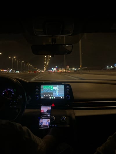 Captured from inside a vehicle, the image presents a night-time scene of driving through a brightly lit city street, suggestive of urban Riyadh, Saudi Arabia. The vehicle's dashboard is visible, featuring a navigation screen displaying a map, a smartphone mounted nearby showing a video, and the steering wheel with illuminated gauges. Through the windshield, a perspective of the road ahead is revealed, displaying a dense array of streetlights, building lights, and taillights of other vehicles indicating traffic. The scene suggests the driver is using navigation while possibly watching content on their phone. The environment is characterized by the intensity of urban illumination at night.