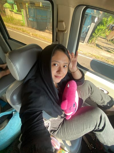 A woman, wearing a black hijab and black long-sleeved shirt, sits in the passenger seat of a vehicle. She is holding a pink stuffed whale toy. Her right hand is raised, with fingers bent. The woman is looking towards the camera. The interior of the vehicle appears to be a van or SUV, with grey leather seats. Outside the window, a road and buildings are visible, suggesting an urban or suburban setting. The lighting indicates it is daytime, with sunlight illuminating the scene. The image is a candid shot, likely taken from another seat within the vehicle. Based on the surrounding environment, the location is Pacing, Indonesia.