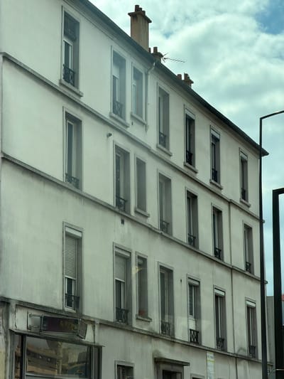 The image shows the side of a multi-story building with a cream-colored facade, angled towards the viewer. The building features numerous rectangular windows, many with dark metal security bars. The windows are arranged in a repeating pattern across the facade, with some appearing to be open. There are also external architectural elements like ledges and a diagonal concrete beam running down the side.
In the upper left of the image, a section of a darker, sloped roof is visible, along with a chimney stack and what appears to be an old television antenna. The sky above is cloudy with patches of blue. Reflections are visible in some of the windows, suggesting the photo might have been taken from across a street or an opposing building. The lighting suggests it could be daytime, possibly overcast.
On the lower right, a larger window displays a reflection of buildings across the way. There is also a small, metallic rectangular object mounted on the wall below this window. A vertical strip of text, appearing to be a label or sign, is visible on the right side of the building, between rows of windows. The overall scene depicts an urban residential or commercial building exterior in Alfortville, France. No people are visible in the image.