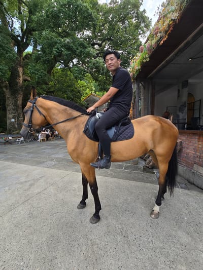 A man in a black t-shirt and black pants is seated on a light brown horse, which is wearing a black saddle and bridle. The man is looking at the camera with a neutral expression. The horse is standing on a paved area in front of a brick building with an overhang decorated with artificial flowers. In the background, large trees and tables with people seated are visible. The scene appears to be outdoors, possibly in the city of Xiaoshan District, China, given the architectural style and vegetation.