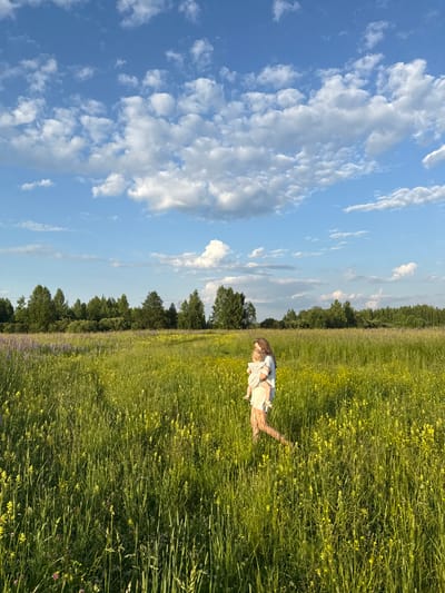 A full-shot, eye-level, outdoor, vertical, daytime photograph captures a woman walking through a lush, grassy field dotted with small yellow flowers. The woman, with long brown hair, is wearing a light-colored dress and is holding a young child, also in light-colored clothing, in her arms. Her legs are visible as she walks forward, with her right foot appearing to be mid-stride. The field is dense with green grass and interspersed with scattered clusters of purple flowers in the foreground and middle ground. In the background, a dense line of trees forms a dark green horizon, against a bright blue sky filled with fluffy white clouds. The lighting suggests it is late afternoon or early evening, with a warm glow illuminating the scene. The location is identified as Koloksha, Russia.