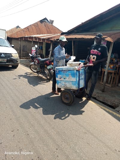 The image shows a street scene in Ilobu, Nigeria (latitude: 7.84, longitude: 4.485). Two men are standing next to a blue and white cooler on wheels. One man is wearing a plaid shirt and a white hat, while the other is wearing a black t-shirt with a graphic design and a black baseball cap. They appear to be interacting with each other, possibly in a transaction. A motorcycle is parked near the curb, and a white pickup truck is partially visible in the background. There are several buildings with corrugated metal roofs in the background. The scene is outdoors, on an asphalt street with a yellow stripe along the edge. The weather is sunny and the shadows are cast to the left.