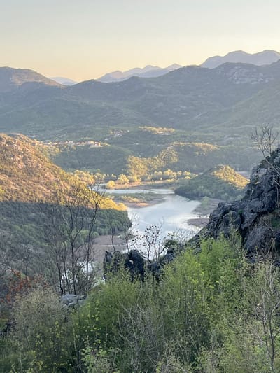 An outdoor landscape photograph depicts a winding river valley in Rvaši, Montenegro. Mountains are seen in the background, with a small village visible along the mountainside. Foliage is prominently featured in the foreground, and the rocky terrain slopes downward towards the river. The scene is illuminated with natural sunlight.