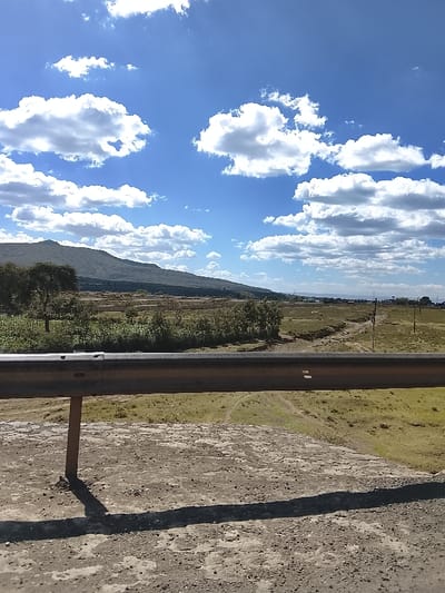 The image depicts a panoramic view of a landscape in Kiambu Township ward, Kenya, under a bright blue sky dotted with numerous white cumulus clouds. In the foreground, a gray concrete surface, possibly a road or bridge deck, occupies the bottom quarter, exhibiting cracks and textured wear. A metal guardrail, dull gray and approximately waist-height, extends horizontally across the lower mid-frame, supported by a single visible vertical post on the left, casting a dark shadow onto the concrete.
Immediately beyond the guardrail, the terrain slopes downward into an expansive, undulating field characterized by patches of dry brown and green vegetation. Multiple dirt paths or unpaved tracks wind through this field. Towards the left midground, clusters of green trees and dense shrubs are visible. Further in the midground, a significant area features rows of partially constructed foundations or unfinished buildings, suggesting an active construction site. Utility poles with overhead lines traverse the right side of the field, extending into the distant landscape.
In the background, a large, dark green mountain range or prominent hills stretch across the horizon from left to center, covered in dense vegetation. The far right horizon displays flatter terrain fading into atmospheric haze. No individuals, overt human actions, or direct interactions are visible within the frame.
