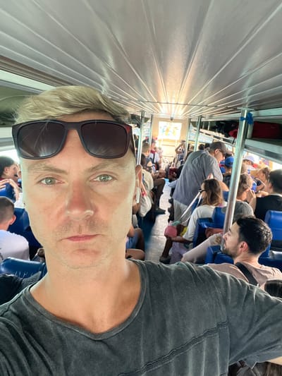 An interior view of a passenger vessel, likely a fast boat or ferry, is depicted, specifically within the harbor area of Toyapakeh, Indonesia. The central subject, a Caucasian male with short blonde hair and light green eyes, approximately 30-50 years old, occupies the foreground, taking a selfie. He wears a dark grey V-neck t-shirt and dark-rimmed sunglasses pushed onto his forehead, with a white earbud in his right ear. His expression is neutral.
The vessel's interior is brightly lit, featuring a light-colored, ridged ceiling and rows of blue passenger seating arranged longitudinally. Numerous passengers of varying ages and ethnicities occupy these seats, indicating a full capacity. In the middle ground, a man in a grey shirt and cap stands while others are seated. A child is visible holding a pink object. Luggage or bags are stored on overhead racks towards the aft section of the vessel. Bright daylight from an open section at the far end of the boat suggests the vessel is either docked or in transit in open waters, consistent with a port departure or arrival.