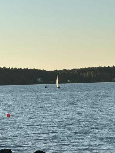 The image depicts a serene lake scene in Solnechnogorsk, Russia, likely during the late afternoon or early evening given the warm, soft light and the gradient sky transitioning from light blue to a pale yellow. The foreground is dominated by the rippling surface of the lake, reflecting the sky and the trees on the distant shore. A red buoy floats on the left side of the lake, marking a section of the water.
In the mid-ground, two sailboats are visible. The larger sailboat, on the right, has a single sail and a lone figure at its helm, cutting gracefully through the water. To its left, a smaller, wider boat carries two individuals, appearing to be paddling or moving slowly.
The background is characterized by a dense line of dark green trees that form the shoreline, suggesting a wooded area surrounding the lake. A few buildings are subtly visible amidst the trees, indicating the presence of human habitation near the water's edge. The overall atmosphere is tranquil and picturesque, capturing a moment of peaceful recreation on the water.