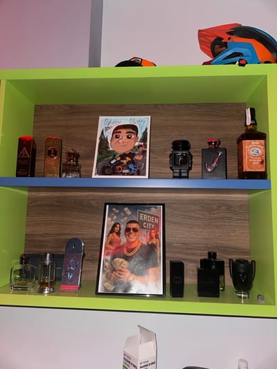 A two-tiered lime green and faux wood-grain shelving unit is displayed against a white wall.

On the top shelf, from left to right, are three perfume bottles: a rectangular gold bottle, a taller rectangular gold bottle, and a clear bottle with a crown-shaped stopper. Next is a vertical white card featuring a cartoon caricature of a male on a black motorcycle, with "Happy B-day" written at the top. Following this are a black robot-shaped figurine, a black rectangular perfume bottle with a red 'V' design, and a brown glass bottle of "Дон Светослав" Bulgarian Whiskey. Above the top shelf, an orange and blue motocross helmet rests alongside a black and orange cap.

On the bottom shelf, from left to right, are two clear glass perfume bottles, followed by a silver elongated object with a blue butterfly design. Centered on this shelf is a framed picture depicting a light-skinned male wearing sunglasses and a gold chain, holding stacks of US dollar bills, with two women positioned behind him. Falling dollar bills and the prominent red text "ERDEN CITY" are visible above the male figure. To the right of the framed picture are three black rectangular perfume bottles, and a chalice-shaped clear glass perfume bottle.

Below the shelving unit, a partially open white cardboard box with "MAGNE D'ORO LIQUID" printed on its side is visible. The prominent text "ERDEN CITY" within the framed picture indicates the city of Erden, Bulgaria.