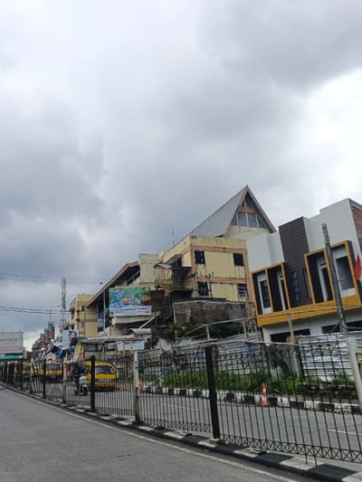 This image depicts an urban street scene in Palayan, Philippines, under an overcast sky. The foreground shows a paved asphalt road with a black metal fence separating it from a raised embankment and adjacent properties. The fence features a decorative cross-hatch pattern on its lower section. A black and white striped curb delineates the road edge. An orange traffic cone is visible on the grassy area behind the fence on the right.
On the left side of the road, several yellow public utility vehicles, consistent with Philippine jeepneys, are visible. One white motorcycle with a rider is positioned on the road, facing left. Utility poles with overhead lines are present along the left side of the street.
The midground features a cluster of buildings. A prominent, multi-story structure on the left has a steep, triangular, grey-tiled roof and light yellow exterior walls exhibiting some wear and darkened window frames. Below and to its right, a large advertising billboard depicting food items is affixed to another building. Further right, a more modern-looking building with white, brown, and yellow facade panels, and rectangular windows, is visible. A red flag is attached to the exterior of this modern building. Between the road fence and the main buildings, there are various smaller, less defined structures and an embankment with some vegetation.
No explicit human interactions are discernible, though the presence of vehicles and structures indicates human activity. The overall environment is that of a bustling urban or commercial area.