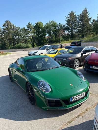 In Arbanassi, Bulgaria, a group of parked cars are visible on a sunny day. The foreground is dominated by a vibrant green Porsche 911. Next to it, a dark gray sedan, a bright yellow car, and a white SUV are parked. Behind these vehicles, a black SUV and a red car are also present. The cars are situated on a paved area, likely a parking lot, surrounded by lush green trees and vegetation. The sky is clear and blue. A man wearing a backpack is standing between the yellow car and the white SUV.