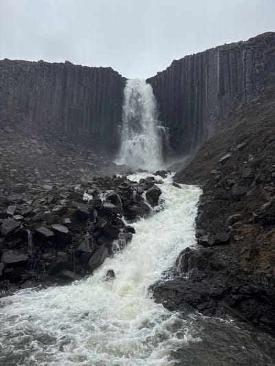 The image, originally rotated 90 degrees clockwise, depicts a powerful waterfall cascading down a dark, rocky cliff face. A prominent feature on the left (when oriented correctly) is a massive cliff displaying distinct horizontal layers and vertical fractures, characteristic of columnar basalt. Sparse, dry vegetation can be seen clinging to the upper sections of this rugged, dark rock formation.

The main body of the waterfall plunges forcefully, creating a wide, frothy, white torrent as it crashes onto a chaotic collection of large, jagged dark rocks at the base of the cliff. To the left of the primary cascade, a smaller, thinner stream of water also descends, partially obscured by the spray and mist generated by the main falls. The turbulent water below is churned into white foam, eventually flowing into a darker body of water, likely a river or pool.

The sky is a uniform, bright overcast grey, indicating a cloudy daytime setting with diffused light. No people, man-made objects, or visible text are present in the scene. The dramatic geological features, particularly the columnar basalt and powerful waterfall, are suggestive of an Icelandic landscape, as hinted by the provided context.