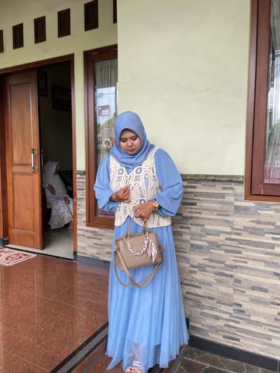An adult female stands outdoors in Ketegan, Indonesia, positioned in front of a residential building. She wears a light blue hijab, a matching light blue long-sleeved dress, and a cream-colored crocheted vest. Her left wrist displays a gold-toned watch, and she holds a light brown handbag adorned with a gold-toned chain detail and a patterned scarf tied to its handle; the bag also has a long shoulder strap. Her gaze is directed downwards towards the handbag. She is wearing light-colored sandals. In the background, visible through an open brown wooden doorway to her left, another individual is seated on a dark chair or sofa, partially obscured and wearing a light-colored hijab and a patterned dress. The building's exterior features a light beige painted wall, a section of textured grey and brown horizontal brick-patterned wall, and two brown wooden window or door frames. Above the main door, four small, dark wooden rectangular accents are present. The ground is covered with glossy reddish-brown tiles featuring dark contrasting borders.