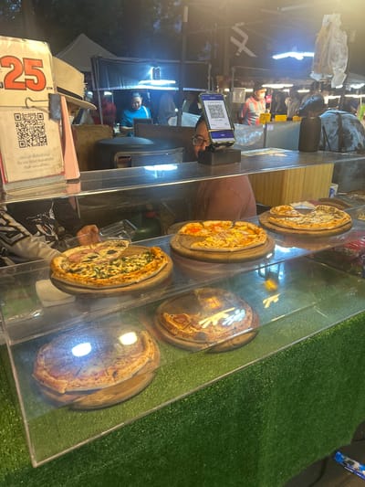 In Tha Khlong Town Municipality, Thailand, a street food vendor is selling pizzas from a stall. The stall is set up with a glass display case showcasing several pizzas on wooden boards. In the foreground, a large pizza with what appears to be spinach and ham is displayed. Behind it, other pizzas with various toppings are visible. A sign with the number "25" in red is prominently displayed on the left, possibly indicating a price. A QR code is also visible on the sign, suggesting a cashless payment option. A person wearing glasses and a dark shirt is partially visible behind the counter, interacting with the display. In the background, other vendors and customers can be seen, and tents suggest a bustling market environment. The overall setting is a nighttime market scene with artificial lighting illuminating the food stall.