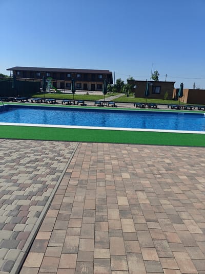 In Shkurupiivka, Ukraine, on a clear, sunny day, an outdoor swimming pool area is depicted. The pool itself is rectangular and filled with vibrant blue water, bordered by a white edge and a strip of artificial green turf. Beyond the turf, a paved walkway constructed from multi-colored interlocking pavers extends into the foreground.
To the left of the pool, several lounge chairs with dark blue cushions are arranged, some partially covered by green umbrellas. Further back, behind the pool and lounge chairs, a long, multi-story building with a dark brown roof and exterior siding is visible. This building has numerous windows and balconies. Adjacent to the building, a children's playground with a slide is present.
To the right of the main building, a smaller, wooden structure with a dark roof and a window can be seen, also shaded by green umbrellas. A wooden fence encloses part of the property in the background. The sky is a uniform, clear blue, with no clouds visible. The scene appears to be a resort or a recreational facility.
