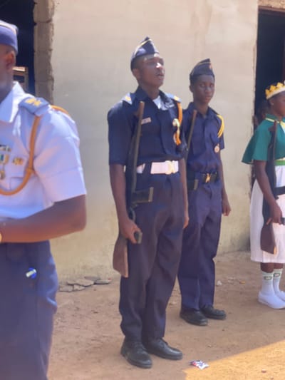 The image, oriented vertically, captures a group of individuals, predominantly young people, standing outdoors in what appears to be a formal or ceremonial setting in Andaha, Nigeria.
In the foreground, partially visible, are two individuals. One wears a white uniform shirt with gold trim on the collar and epaulets, a dark tie, and a dark cap with a gold emblem. The other, mostly out of frame, wears a dark blue uniform.
Behind them stand two young men in full dark blue uniforms, complete with gold-trimmed epaulets and dark blue caps with emblems. They both hold long, slender wooden staffs or mock rifles, positioned vertically at their sides. They are wearing black shoes. On the light brown, dirt ground near their feet, a crumpled piece of litter, possibly a wrapper, is visible.
Further back and slightly elevated, a young woman is seen. She is dressed in a white gown or dress, adorned with a green sash, and wears a distinctive yellow and white patterned head covering, resembling a crown. She has white socks with green patterns and white shoes. She appears to be holding a dark brown object.
The background consists of a plain, light-colored wall, possibly beige or light brown, suggesting an architectural structure. Above the wall on the left, a darker, shadowed area might indicate a window or opening. The lighting suggests it is daytime, with bright sunlight illuminating the scene, indicative of clear weather. The individuals' expressions appear focused or neutral, consistent with participation in a formal event, possibly a parade or youth group gathering. No discernible text is visible in the image.