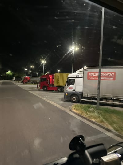 A nighttime scene captures several parked semi-trucks on a paved lot, likely a rest stop or truck yard, located in Luckau, Germany. The foreground is dominated by the interior of a vehicle, with a black dashboard and steering wheel visible. In the midground, multiple semi-trucks are lined up. The closest truck is white with a large grey trailer bearing the red logo "GRABOWSKI FURNITURE LOGISTICS." To its left are two red semi-trucks, one with a yellow container. Further down the line, another white truck is visible. The background consists of streetlights illuminating the area, trees, and a dark sky. The ground is asphalt, with some patches of grass along the edge.