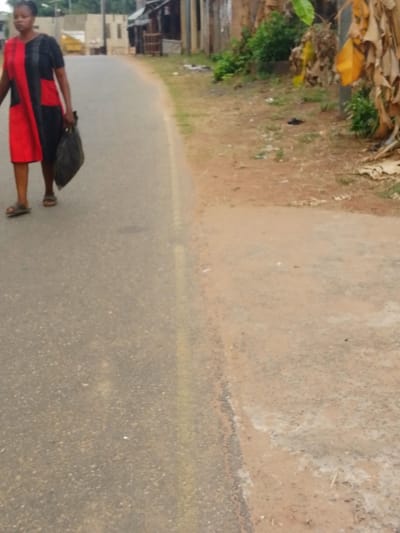 A female individual is observed walking from left to right along a paved asphalt road in Umuoji, Nigeria. She wears a knee-length dress, split vertically with a red section on her left side and a black section on her right side, and dark sandals. A dark-colored plastic bag is carried in her right hand, hanging by her side. The road features a faded single yellow line running parallel to the right edge. To the right of the paved road is an unpaved dirt shoulder composed of reddish-brown soil, sparse green vegetation, and scattered dry leaves. Further right, a large plant with numerous dry, yellow, and brown leaves is visible. In the background, along the left side of the road, several low-rise buildings constructed from various materials are present.