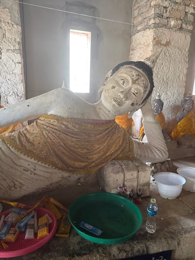 This image depicts a large, reclining Buddha statue, likely made of plaster or concrete, within a partially constructed building. The statue is adorned with a golden, sequined robe and has what appears to be gold leaf applied to its face and body. To the right of the reclining Buddha, three smaller, dark-colored Buddha statues are visible, also draped in yellow fabric.
In the foreground, there are several objects placed on the ground. A pink plastic basket contains various candles, some in packaging and others loose. Next to it is a green plastic tray, and a plastic bottle of water. Behind the green tray, two small decorative figurines and some artificial flowers are placed near the base of the reclining Buddha. Two white plastic bowls are also visible in the lower right corner.
The environment suggests an ongoing construction or restoration project. The walls are a mix of rough stone and smoother plaster, with exposed bricks in some areas. A rectangular window, emitting bright light, is positioned in the background on the left.
The location is identified as Baan Harn, Thailand.