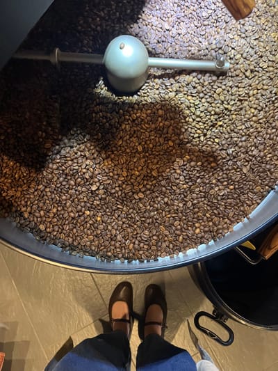 The image displays an overhead perspective within a coffee processing facility located in Meisenheim, Germany. The primary focus is a large, circular, metal cooling tray filled with a substantial quantity of roasted coffee beans, exhibiting various shades of brown. A metallic agitator mechanism, consisting of a central spherical component and two horizontal bars, is positioned within the coffee beans, casting a shadow across them. In the foreground, the lower legs and feet of an individual are visible, clad in dark trousers and dark Mary Jane shoes with a strap across the instep. The individual stands on a tiled floor composed of light-colored, square tiles. To the lower right, a portion of a black cylindrical container with a handle is visible on the floor. The scene is illuminated by overhead lighting, creating defined shadows.