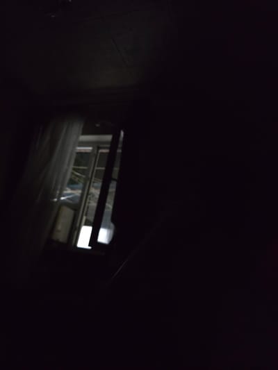 An underexposed image taken from indoors looking out of a window. The interior is mostly dark, with a pale, sheer curtain visible on the left side of the frame. The window shows a view of what appears to be an outdoor area, possibly a courtyard or backyard, with some indistinct shapes and structures. There is a bright rectangular light source visible through the window, suggesting a light or a brightly lit area outside. The overall impression is of a dimly lit room at night or in low-light conditions, with a view to the outside. There is no discernible location information in the image.