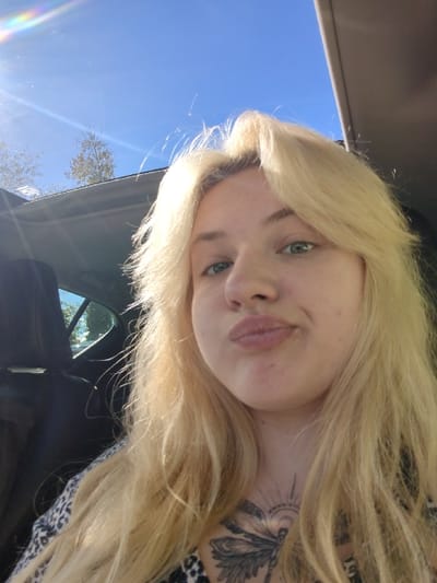 A female individual with long, light blonde hair and light green eyes is positioned in the foreground, facing the camera. She wears a patterned top and displays a large tattoo on her upper sternum featuring a winged insect and a sunburst design. Her lips are pursed in a pouting or kissing expression. The scene is set inside a vehicle, with a dark headrest visible to her left. Above and to her left, an open sunroof or window reveals a bright blue sky with a prominent sun flare emanating from the upper left corner. Silhouettes of trees with light foliage are visible against the sky. The specific location within Maple Ridge, Canada, cannot be determined from the visual evidence provided.