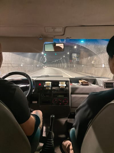 The image shows the interior of a van driving through a tunnel. The viewpoint is from the passenger seat looking forward, with the driver's back visible. The dashboard features a steering wheel, a gear shift, and various controls. Two paper cups are in a holder on the dashboard. A wallet and other items are placed on the dashboard in front of the passenger seat. The tunnel walls are visible, illuminated by overhead lights, and the road ahead shows other vehicles. The scene appears to be nighttime, and the van is in motion. The overall impression is one of travel. The location is identified as the city of Gorno Kamartsi, Bulgaria, and the tunnel is likely part of a road network within or near the city.