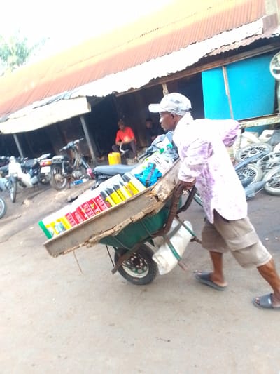 In Orlu, Nigeria, an individual wearing a patterned shirt, cap, and khaki shorts is pushing a wheelbarrow along a paved surface. The wheelbarrow is filled with various items, possibly goods for sale. In the background, there are several motorcycles parked near a building with a corrugated metal roof. Another person is seated inside the building. Additional spare motorcycle parts are stacked on the side of the building. The environment is likely a market area or a roadside commercial zone.