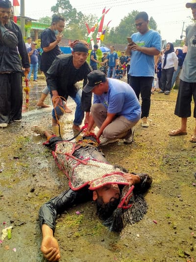 A man lies in muddy water wearing a traditional outfit with a head ornament and long hair. Two men, one with a red headband and another in a cap, attend to him, possibly untying ropes around his body. The scene is surrounded by onlookers, some holding phones, suggesting a public display or performance. The setting includes banners and what appears to be a village or rural environment.