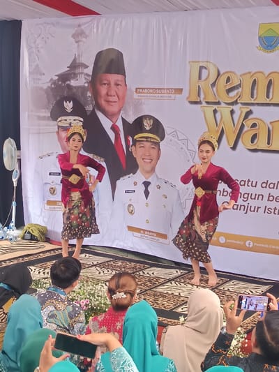 The image depicts a cultural performance in Kawungluwuk, Cianjur Regency, Indonesia. Two young women, barefoot and dressed in traditional attire consisting of dark red velvet kebayas, patterned batik skirts, and ornate golden headpieces, are performing a dance on a stage covered with a patterned carpet. Their postures feature bent arms and precise hand positions. Behind them, a large white banner serves as a backdrop, displaying portraits of political figures: Prabowo Subianto, identified as "PRESIDEN REPUBLIK INDONESIA," is prominently centered. To his right, a smiling Dr. Wahyu is identified as "BUPATI CIANJUR" (Regent of Cianjur), and a "GUBERNUR" (Governor) is partially visible to Prabowo's left. The banner also incorporates architectural imagery resembling the Cianjur Great Mosque and text, including references to "PEMKAB CIANJUR" and "Cianjur Istimewa." In the foreground, an audience, some wearing hijabs, observes the performance from behind, with at least two individuals actively recording the event using smartphones. A white pedestal fan and plastic water bottles are positioned on the left side of the stage, which is further adorned with white floral arrangements along its front edge.