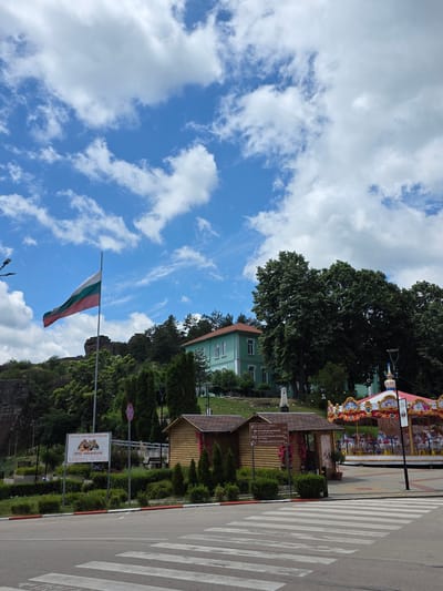 The image captures a scene in the city of Belogradchik, Bulgaria, featuring a large Bulgarian flag waving in the foreground against a backdrop of a bright blue sky with scattered white clouds. To the left of the flag is a rocky cliff formation, and further to the right is a light green building with a red-tiled roof, likely a public building. In the mid-ground, there are several small wooden structures, one of which appears to be a kiosk or information booth, and a colorful carousel. The scene also includes trees, bushes, and a street with a pedestrian crossing in the lower part of the image. A sign with Bulgarian text and the words "DIRTY ADVENTURE" is visible on the left side. The overall impression is a public space, possibly a park or town square, during daytime.