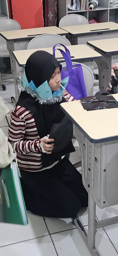 A young female wearing a black hijab and a striped long-sleeved shirt is seated on the floor in a classroom in Bekasi, Indonesia. She has a light blue, heart-patterned paper crown with a silver tinsel trim around her neck. She is holding a black object, possibly a hat or a bag, in her hands and has a lollipop in her mouth. The classroom features desks with light wood tops and white metal frames. A purple tote bag is visible on a desk behind her. The floor is tiled with a white and grey grid pattern.