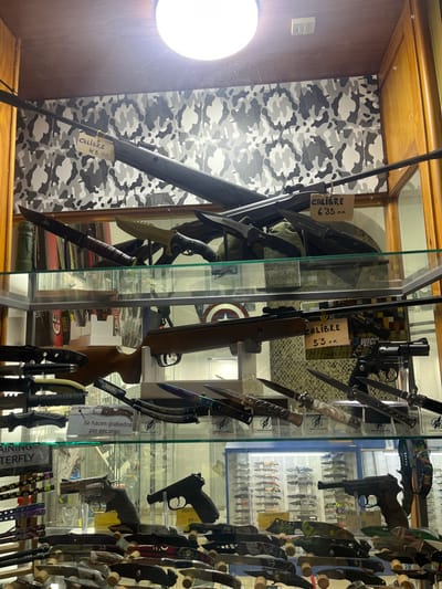 The image displays a multi-tiered glass and wooden retail display case in a store located in Ayamonte, Spain, showcasing various weapons and knives against a grey and black camouflage-patterned background wall.
**Top Shelf:** Two long-barreled rifles or air rifles are angled across the upper section. The top-most rifle, black, features a tag indicating "CALIBRE 4.5 MM". Below it, another black rifle is marked with a tag reading "CALIBRE 6.35 MM". Beneath these rifles, several large fixed-blade knives are positioned, including a dark combat knife on the left, a tan-bladed combat knife, and multiple dark-bladed knives, some labeled "PREDATOR".
**Middle Shelf:** A wooden-stock rifle or air rifle is horizontally presented, with a tag stating "CALIBRE 5.5 MM". To its right, a black revolver with a dark grip stands upright. Various folding and fixed-blade knives are arranged in individual clear stands, including those with iridescent blades and an ornate switchblade-style knife featuring a light-colored handle. A small white sign, "Se hacen grabados por encargo." (Engravings are done by order.), is visible. A Captain America shield emblem and a "WICK" logo are faintly discernible in the background.
**Bottom Shelf:** This shelf contains several handguns: a silver-colored revolver with a dark grip on the left, a black semi-automatic pistol centrally located, and a second semi-automatic pistol, possibly a Luger P08 replica, on the right. Numerous folding knives, multi-tools, and small blades are extensively displayed horizontally on stands. A sign on the left edge reads "TRAINING BUTTERFLY". In the background, a blue-framed display case filled with additional small knives is visible.
The display is illuminated by a central circular overhead light fixture. No individuals are present in the scene.