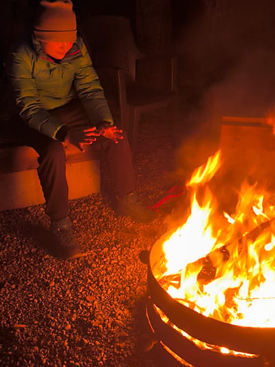 A person in a light green jacket and a brown beanie sits on a wooden bench next to a fire pit. The person is warming their hands over the fire. The fire pit is made of metal and contains burning wood. The ground around the fire pit is covered in small stones. Two chairs are behind the seated person. Based on geocoding data, the location is Lilaste, Latvia.