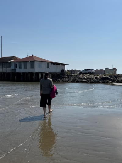 A person is wading in the water at the beach in Anzio, Italy. The person is wearing a gray top and black shorts, and is carrying a pink backpack. The beach is sandy and wet, and there is a building on stilts on the left. In the distance, there are buildings and a car on a rocky breakwater. The sky is blue.