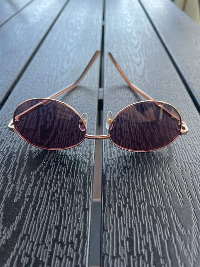 The image shows a pair of sunglasses resting on a dark, textured wooden surface, likely a table, in Zaķumuiža, Latvia. The sunglasses feature round or oval dark-tinted lenses and thin, gold or rose gold-toned metallic frames and arms. The surface consists of horizontal planks with a prominent, embossed wood grain pattern, appearing to be stained or painted a deep, matte black. Bright, natural light illuminates the scene, casting reflections on the sunglass lenses and accentuating the textured details of the table. The strong lighting suggests it is daytime, possibly sunny. There is no discernible activity taking place, and no people are visible in the frame. No text is visible in the image.