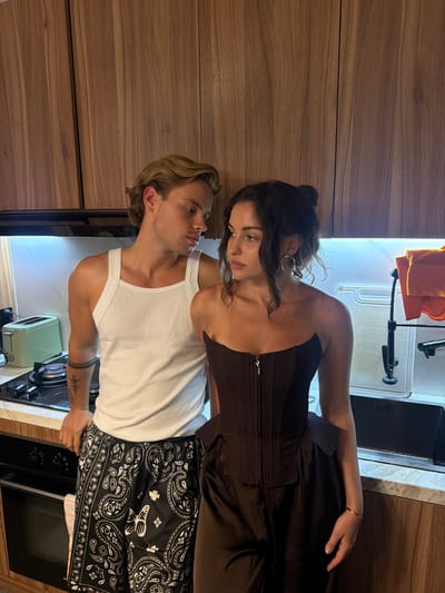 A woman and a man are standing close to each other in a kitchen. The woman has dark hair pulled into a bun and is wearing a brown corset top. The man has blond hair and is wearing a white tank top and black bandana-patterned shorts. They are both looking at each other.
The kitchen has wooden cabinets, a white countertop, and a stainless steel sink. There is a toaster oven on the counter next to the man.
The location is Kerobokan, Indonesia.