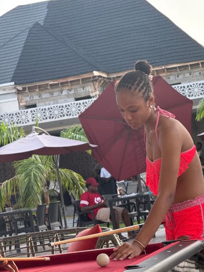 A medium outdoor shot shows a young woman playing pool. She is wearing a pink bathing suit and is leaning over a red pool table, cue stick in hand, poised to strike a white ball. The background shows other people seated, palm trees, umbrellas, and a multi-story building, typical of Rubuchi, Nigeria.