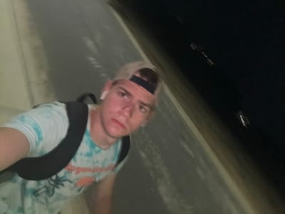 A young man wearing a tie-dye t-shirt and a baseball cap is taking a selfie while walking down a paved path at night in Panama City Beach, United States of America. He has white wireless earbuds in his ears and is carrying a black backpack. The path is bordered by a light-colored wall on the left and a dark, out-of-focus area on the right, possibly vegetation or the night sky. There are a few blurry lights visible in the dark area to the right. The man's face is illuminated, suggesting a light source from the front.