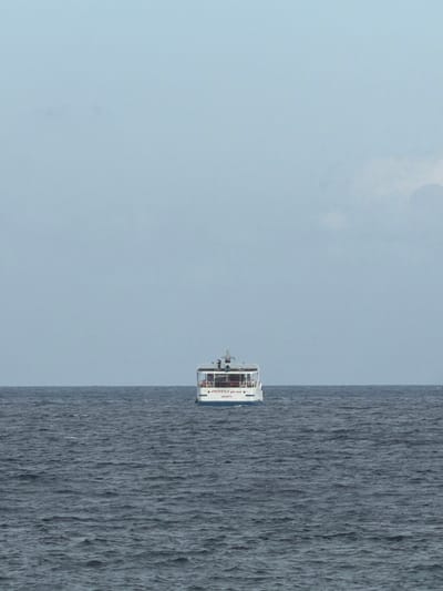 A small, white passenger boat with a red stripe and "JAKARTA" written on its stern is sailing on a dark, choppy ocean under a pale blue, overcast sky. The boat is positioned centrally in the frame, appearing distant. The sea occupies the lower half of the image, while the sky fills the upper half. There are no people visible on the boat, nor any other objects or structures in the immediate vicinity. The scene suggests a calm, maritime environment likely near the coast of Manggis, Indonesia, given the mention of "JAKARTA" on the boat, which is a major port city in Indonesia.