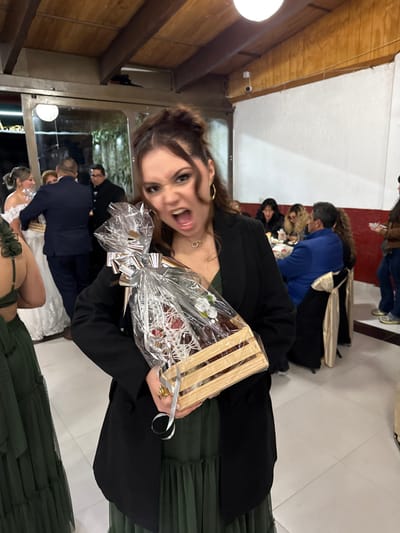 A woman poses, sticking out her tongue and holding a wrapped gift basket. She wears a black blazer, green dress, and gold hoop earrings. Behind her, blurred figures stand on white tiled floors. A wedding scene is visible through a mirrored wall. Other people sit at tables in the background. A light fixture is visible in the upper left corner.