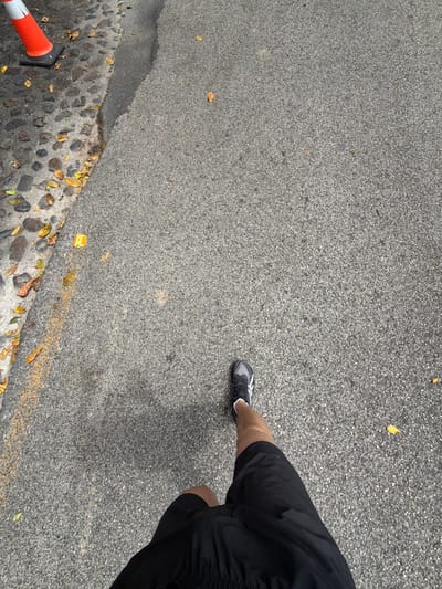 The image displays a first-person, downward-angled view of a person's lower body in La Romana, Dominican Republic. The subject is wearing black athletic shorts and a black and white athletic shoe on the visible left foot, which is positioned on a grey, granular asphalt surface. To the left, the asphalt transitions into a darker, irregularly textured area composed of cobblestones or small, rough stones. A faded yellow painted line runs along the edge of the asphalt adjacent to the cobbled section. Scattered yellowish-brown fallen leaves are visible on both the cobblestones and the asphalt. In the upper-left corner of the frame, a partial view of an orange traffic cone with a white reflective band is visible, situated on the cobbled surface. Shadows cast across the asphalt indicate the presence of an overhead light source.
