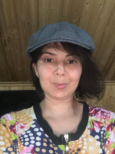 The image shows a medium shot of a woman wearing a grey flat cap and a colorful, patterned shirt with a zipper. She has dark hair and is looking directly at the camera with a slight smile. The background consists of wooden paneled walls. The overall impression is that of a casual portrait, likely taken indoors. There is no information or visual cue to suggest this photo was taken in Kalininskaya, Russia.