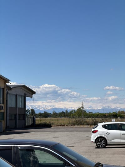 The image is taken in Gudo Visconti, Italy. A two-story building is in the foreground. Beyond the building, there is a view of mountains with snow-capped peaks. Clouds fill the space above the mountains. A fence and some trees are situated between the building and the mountains. A white car is parked on the right side of the image, and a black car is also partially visible in the foreground on the right. The sky is blue.
