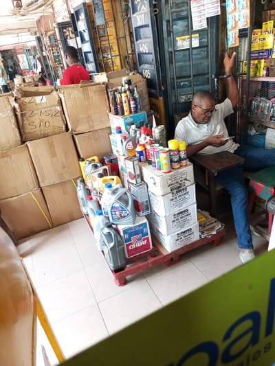 The image captures the interior of a bustling auto parts or general goods shop, likely a market stall, located in Satellite Town, Nigeria. The scene is brightly lit, indicating daytime.
In the foreground, a man wearing glasses and a light-colored, patterned polo shirt is seated on a wooden stool, engrossed in his mobile phone. His left arm is casually raised, resting on the top of a tall metal container that forms part of the shop's structure. His attention is focused downward on the device, suggesting a moment of leisure or waiting.
The shop is filled with a dense array of merchandise. To the left, numerous brown cardboard boxes are stacked high, some handwritten with labels such as "215,2" and "STD". In the center, a red plastic pallet holds an organized display of various automotive products, primarily large plastic containers of motor oil and oil treatments, including brands like "ABRO Motor Oil 20W-50" and "ABRO 500 OIL TREATMENT." Numerous spray cans and smaller bottles of lubricants and additives are also visible on top of cardboard boxes.
The background features robust metal containers serving as the shop's walls, with numbers like "307," "406," and "605" painted on them. One container also bears the word "LAGUNA". Shelves lining these walls are packed with other products, including "TCM EPOXY STEEL GUM" and "GRINDING PASTE". In the left background, another man in a red shirt is partially visible amidst further stacks of boxes, and a third person is faintly seen further in the distance. An old fan hangs from the ceiling. The overall impression is that of a functional, somewhat cluttered, yet organized commercial space.