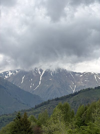 The image depicts a mountainous landscape, possibly near Рилски манастир, Bulgaria, given the prompt's location reference. The mountain slopes are partially covered with snow, especially at higher altitudes. The right side of the image features a forested area, characterized by green vegetation and trees. On the left side, dense clouds obscure the view, suggesting a cloudy or overcast weather condition.