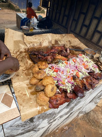 The image shows a close-up, overhead view of a large platter of grilled meats and fried snacks, served on brown paper. The platter is piled high with various cuts of meat, some appearing to be ribs and larger portions of dark, grilled meat. The meat is garnished with shredded cabbage, sliced red onions, and chopped tomatoes. To the side of the meat are several golden-brown, fried balls, possibly plantain fritters or yam balls.
In the foreground, a pair of dark-skinned hands are visible, with one hand reaching into a clear plastic container filled with more of the fried balls. A silver ring is visible on one of the fingers. The hands suggest that someone is in the process of serving or selecting food from the platter.
The setting appears to be an outdoor food stall or market. The background includes elements of a rustic structure with a corrugated metal roof and wooden beams, as well as a partially visible chain-link fence. To the left, a person wearing an orange shirt is visible, along with some bottles of liquid (one clear, one yellow) and a metal bowl. The ground is a mix of dirt and tiled surfaces. The lighting suggests it is daytime, possibly with overcast skies as there are no strong shadows.
The overall impression is of a vibrant, informal food service, likely in a bustling environment. The food looks appetizing and generously portioned. No visible text is present in the image. The location context indicates this scene is from Barakin, Nigeria.